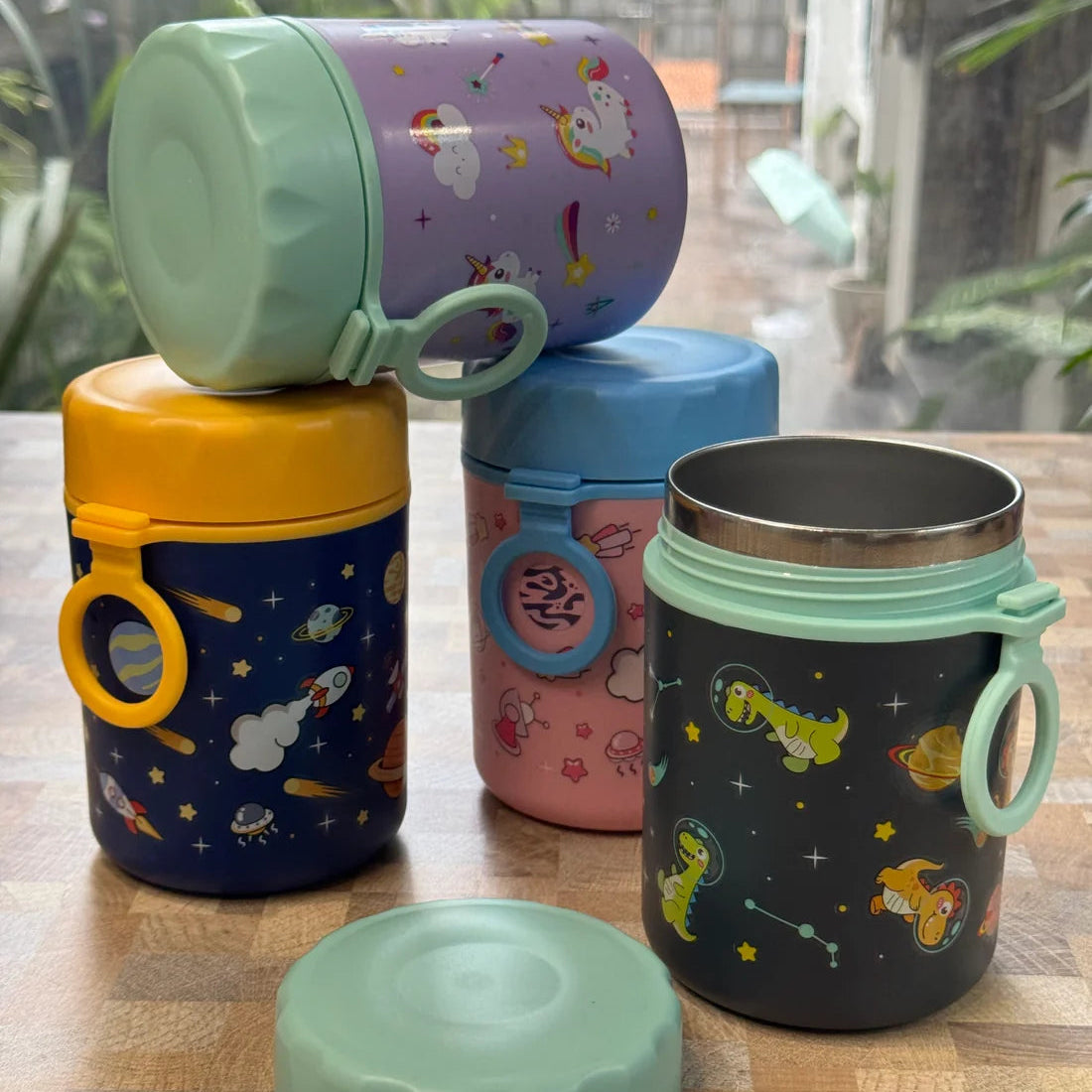 Five reusable insulated tumblers with lids are arranged on a wooden tabletop in front of a window with greenery outside. The tumblers have colorful space-themed prints, including planets, stars, rockets, and astronauts, and each has a loop handle.