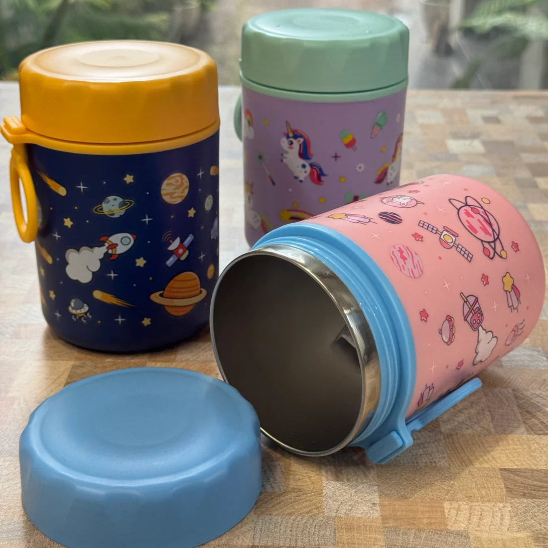 Three insulated stainless steel lunch boxes with colorful cartoon prints are arranged on a wooden tabletop in front of a window. One box is standing upright with a yellow lid and blue handle, one is standing upright with a mint green lid and purple unicorn print, and one is lying on its side with a blue lid placed in front.